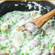 Creamed potatoes and peas in a skillet being stirred with a wooden spoon.